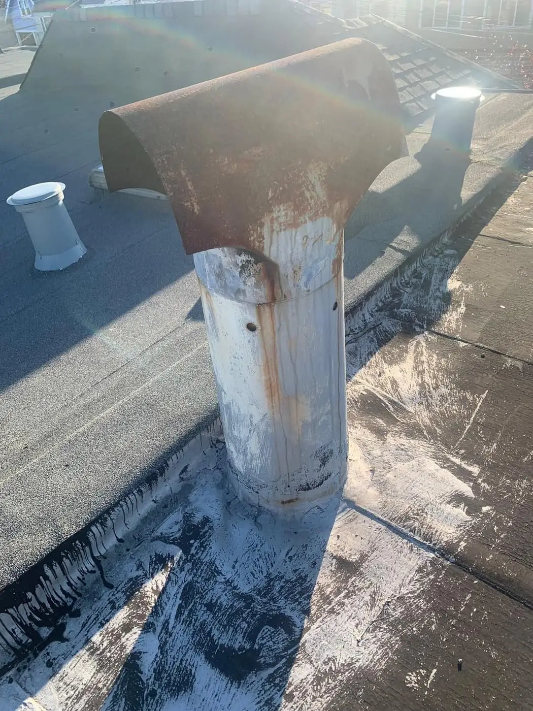 A white metal flue pipe with a rusted hood on a flat roof, with two small roof vents behind it.